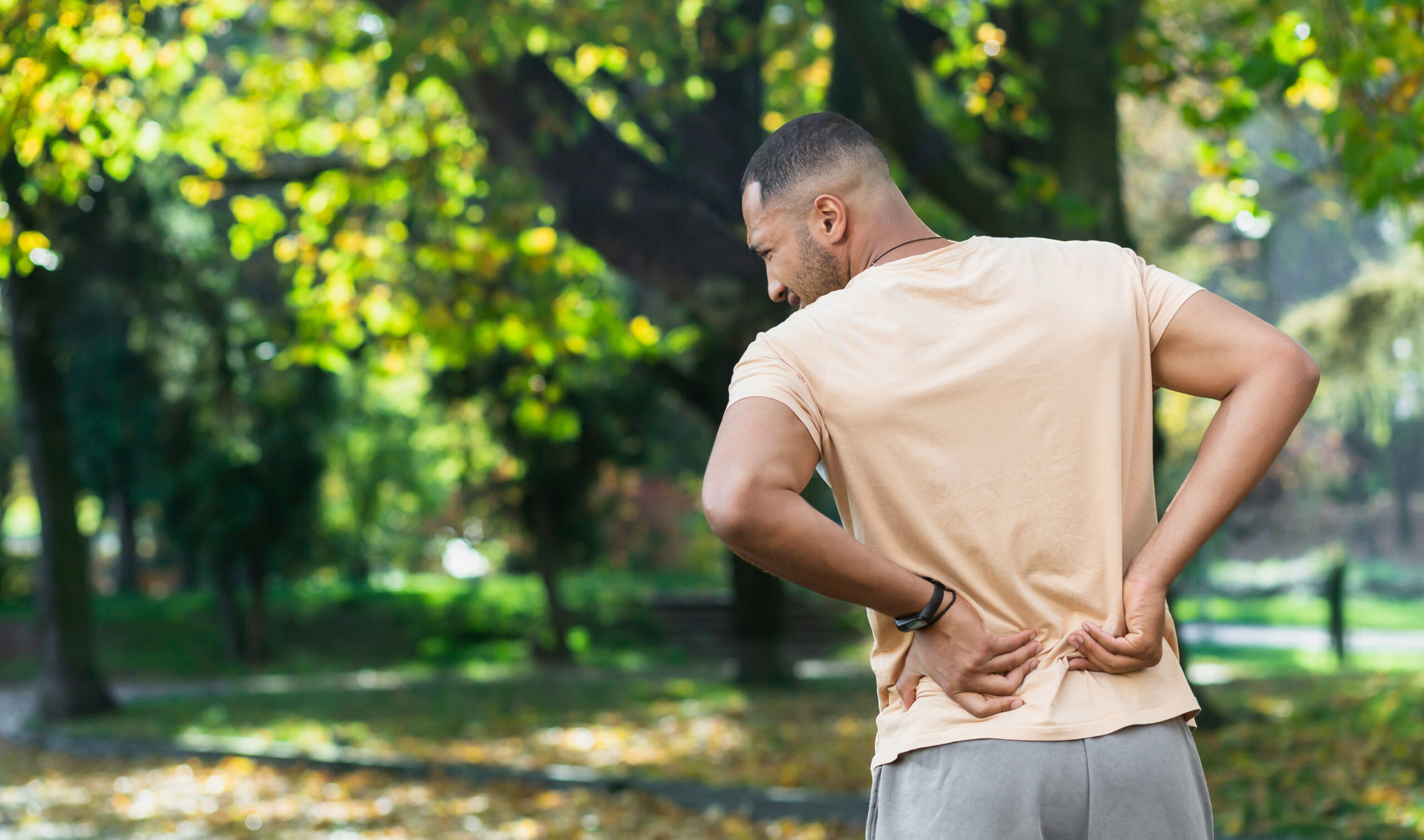 A man standing outside with his back to us, rubbing his lower back and hip area, clearly in pain