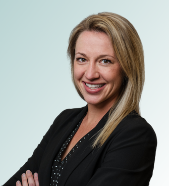 A woman with shoulder-length blonde hair is smiling at the camera. She is wearing a black blazer over a black blouse with small white polka dots. Her arms are crossed, and the background is a light gradient from teal to white, reminiscent of the professional portraits often taken by Jeffrey Ebel, DO.