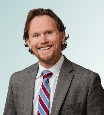 Dr. Dana Clark, MD, with medium-length wavy brown hair and a beard, is smiling warmly. He is wearing a grey checkered suit jacket, a white dress shirt, and a striped blue, red, and white tie. The background features a light gradient of blue.