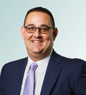 Jeffrey Ebel, dressed in a dark blue suit, white shirt, and light purple tie, is smiling warmly. He has short dark hair and is wearing glasses. The background features a light gradient blue that complements his professional appearance.