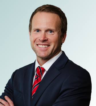 A man with short brown hair, wearing a dark suit, white dress shirt, and red striped tie, smiles at the camera with his arms crossed. The background is a light gradient. Dr. Dana Clark exudes professionalism and confidence in this portrait.