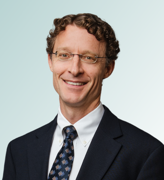 A man with curly hair and glasses, possibly Dr. Jeffrey Ebel, is wearing a dark suit, white shirt, and patterned tie. He is smiling and standing against a light blue gradient background.