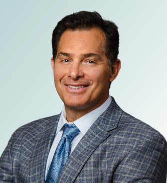 A man with short dark hair, wearing a grey checked suit, a light blue dress shirt, and a blue patterned tie, smiling at the camera against a light gradient background. Dr. Dana Clark, MD, exudes confidence and professionalism.