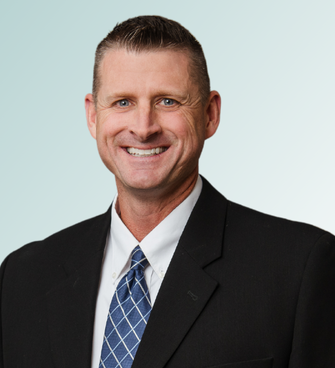 Thomas Anderson, wearing a black suit jacket, white dress shirt, and blue tie with a white checkered pattern, is smiling and has short, neatly-combed hair. The background features a light blue gradient.