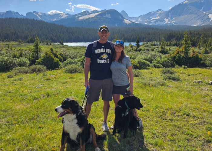 a man and a woman standing on green grass in front of a mountain with two dogs