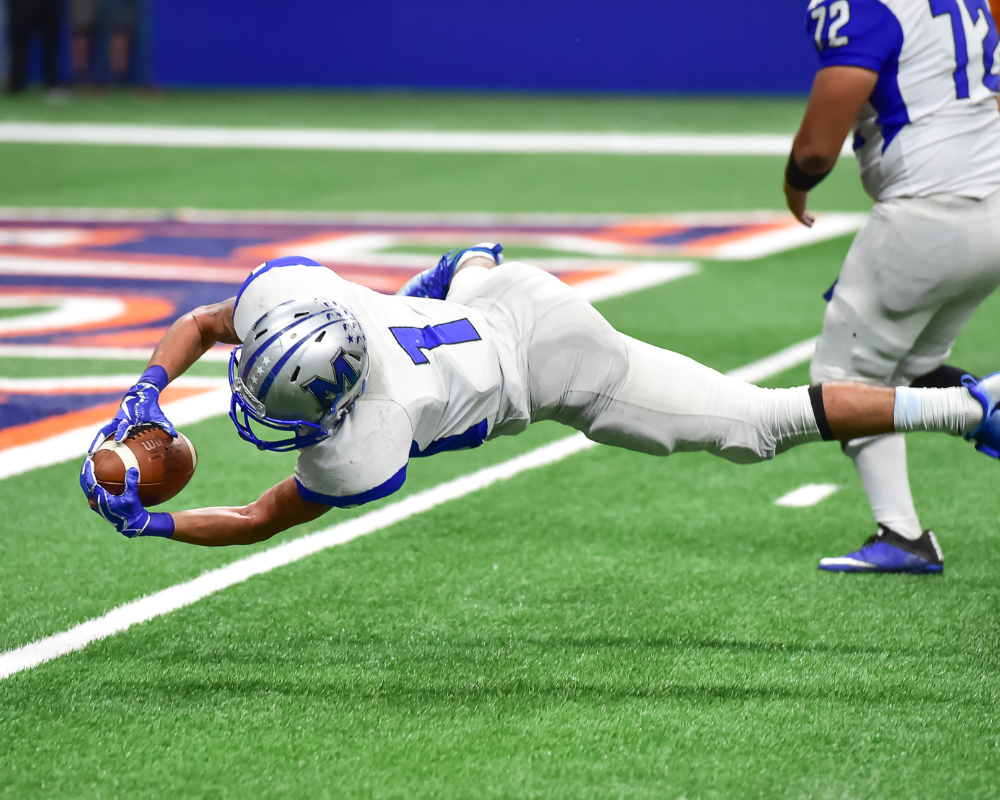 A football player in a white and blue uniform dives forward with an outstretched arm to make a catch. He is mid-air on a green turf field with another player visible in the background. His fingers are just about to grasp the football.