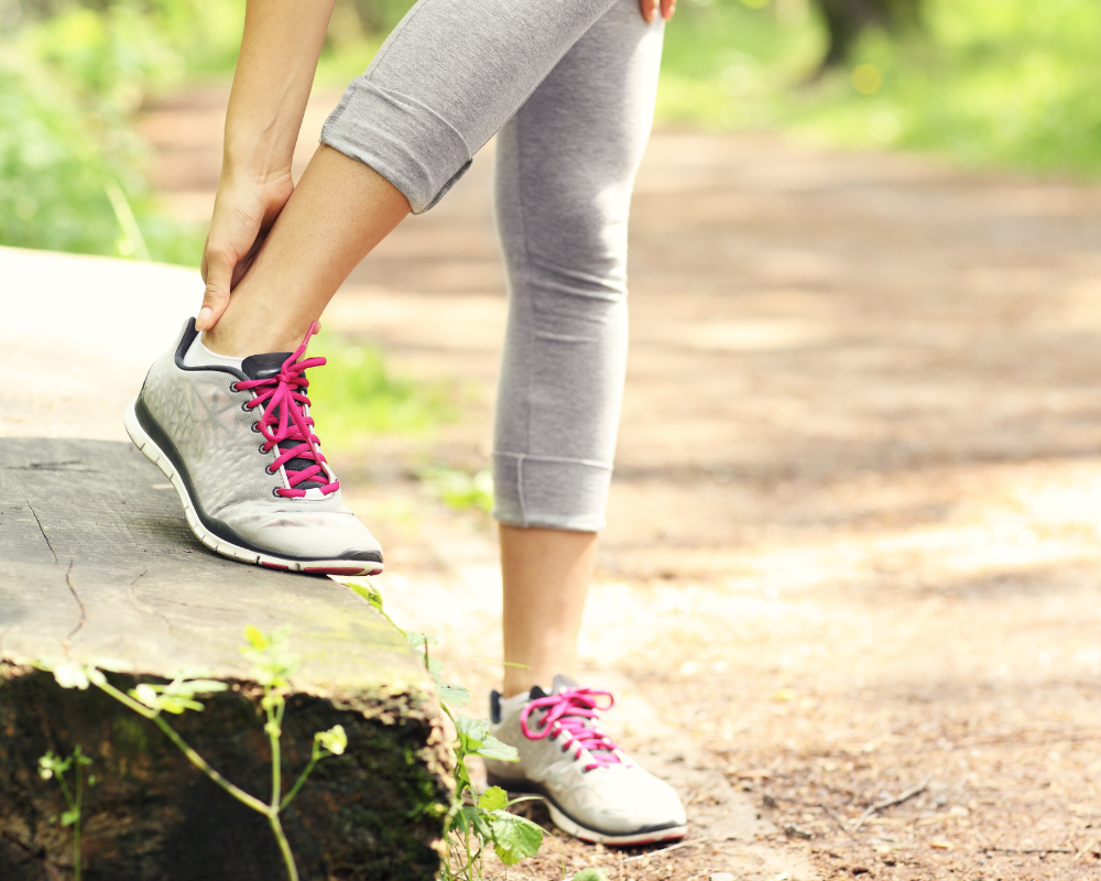 A person in gray athletic leggings and sneakers with pink laces, using one leg to rest on a tree stump while stretching their calf muscle on a wooded trail. The background shows a dirt path lined with greenery and dappled sunlight.