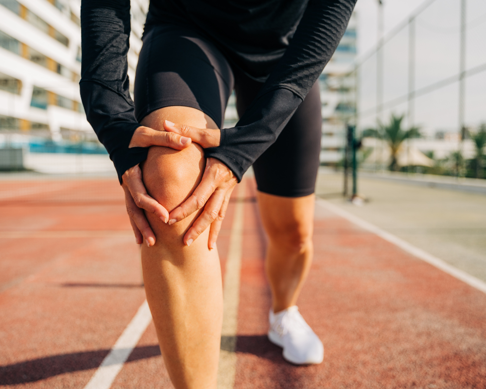 A person in athletic attire is holding their knee in apparent discomfort while standing on a track. The background shows a fence and out-of-focus buildings. The person's face is not visible.