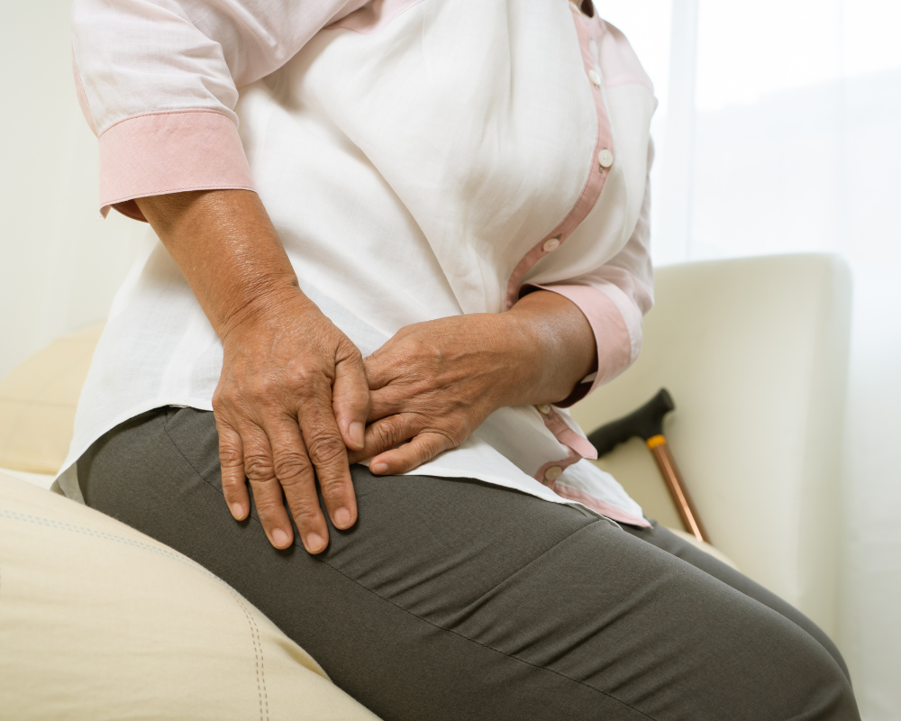 A person wearing a white and pink shirt and gray pants sits on a cushion while clutching their right hip with both hands, suggesting discomfort or pain. A cane rests beside them.