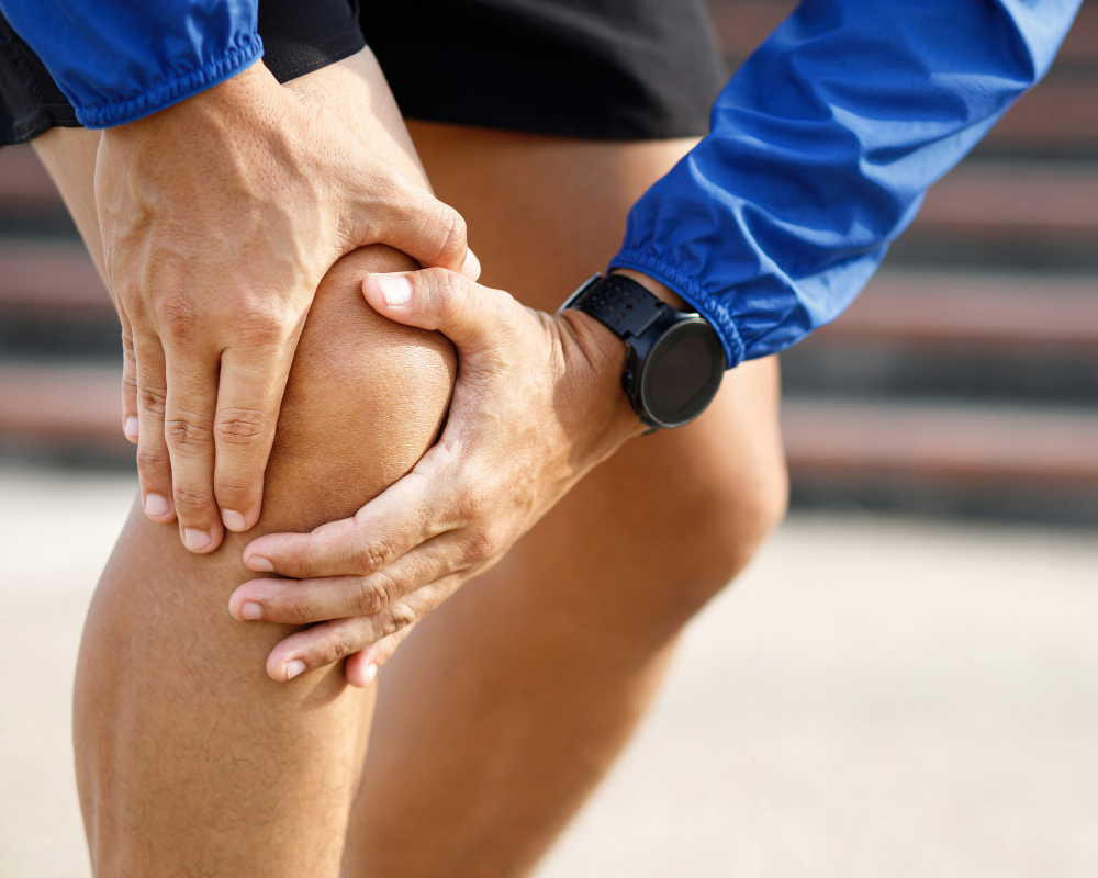 A person wearing a blue long-sleeve shirt and black shorts is holding their right knee with both hands, indicating potential pain or injury. They are wearing a black watch on their left wrist. The background is blurred, suggesting an outdoor setting.