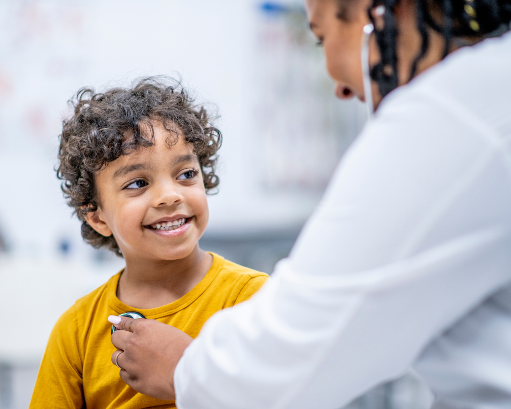 A smiling child with curly hair wearing a yellow shirt is sitting in a medical office while a healthcare professional in a white coat uses a stethoscope to check their heartbeat. The child looks happy and comfortable during the examination.