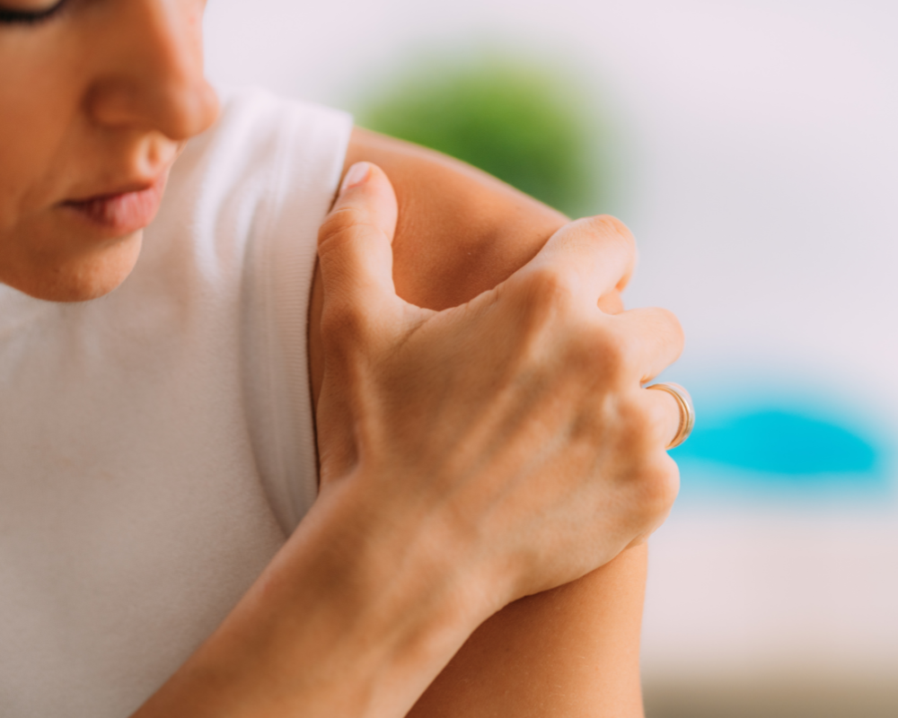 A person wearing a white sleeveless top is touching their right shoulder with their left hand. The focus is on their shoulder and hand, showing a ring on their left ring finger. The background is blurry with a touch of greenery and blue hues.