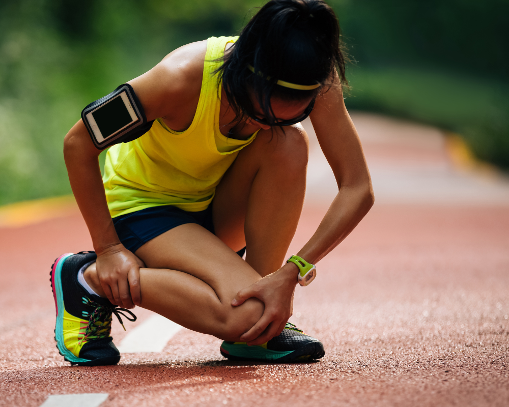 A person dressed in athletic gear crouches down on a running track, holding their lower leg with a pained expression. They are wearing a yellow tank top, dark shorts, colorful running shoes, a fitness tracker, and an armband for a device.