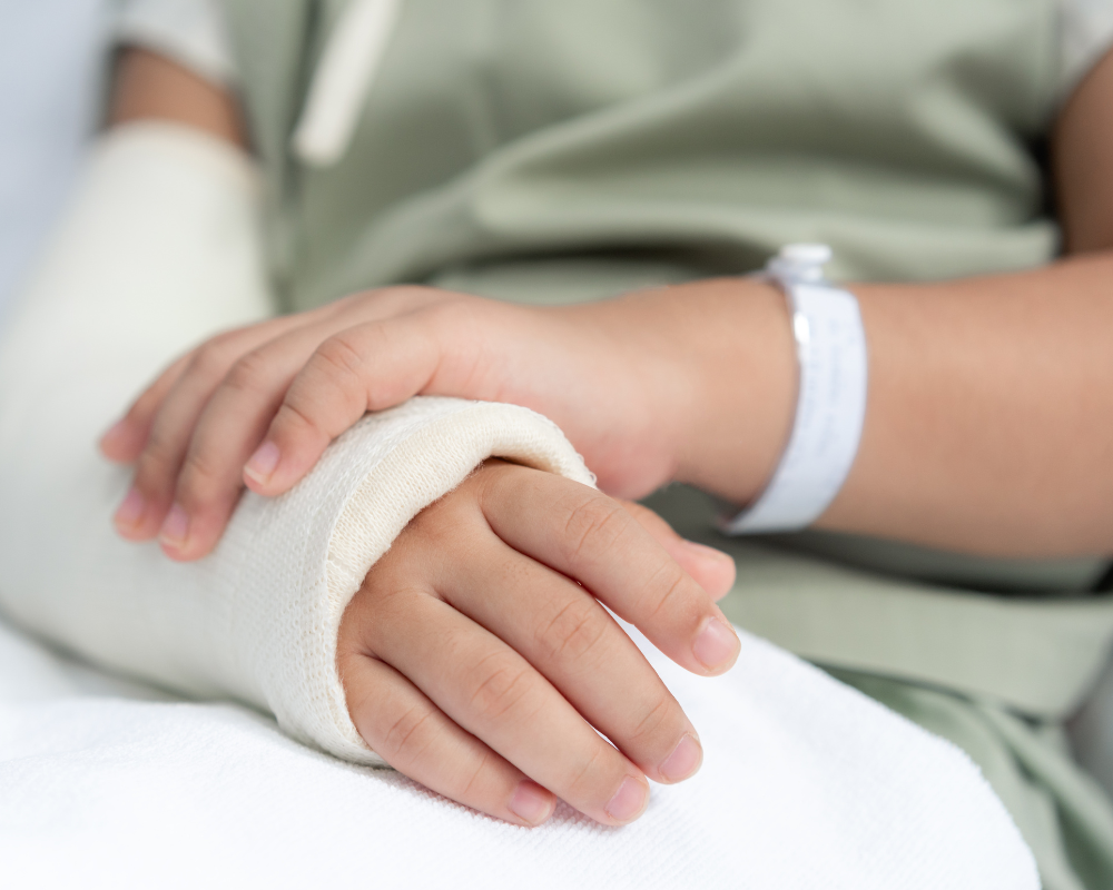 A close-up of a person's hands resting on their lap. The left hand is wrapped in a white bandage, and the person is wearing a hospital wristband. The individual is dressed in a light green hospital gown and is sitting on a white sheet.