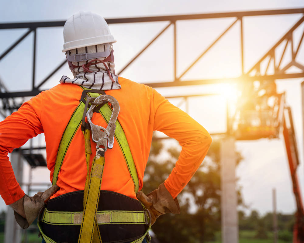 A construction worker in an orange shirt, white helmet, and safety harness stands with hands on hips, looking at a metal framework structure being built. The sun is setting in the background, casting a warm glow over the construction site.