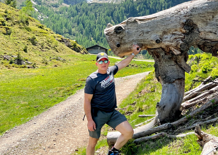 A person wearing a cap and sunglasses is standing on a trail in a mountainous area. They are holding onto a large, uniquely-shaped tree trunk with one hand. 