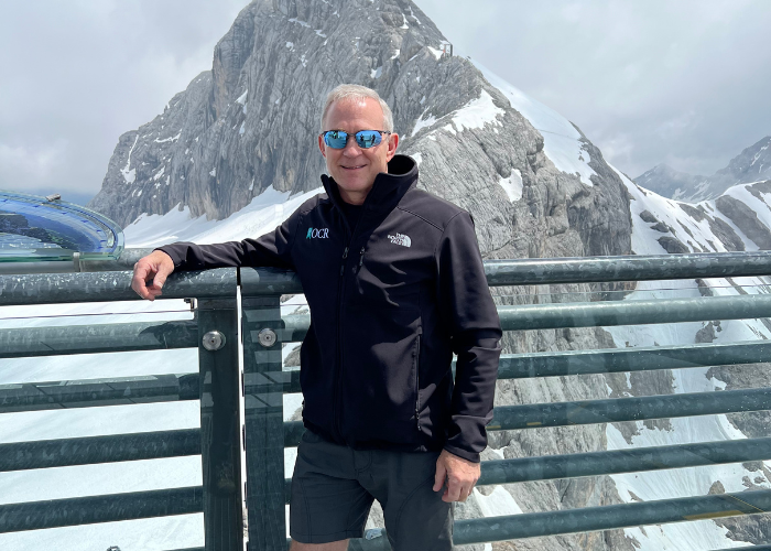 A man wearing sunglasses, a black jacket, and shorts stands by a metal railing with snow-covered mountains in the background. 