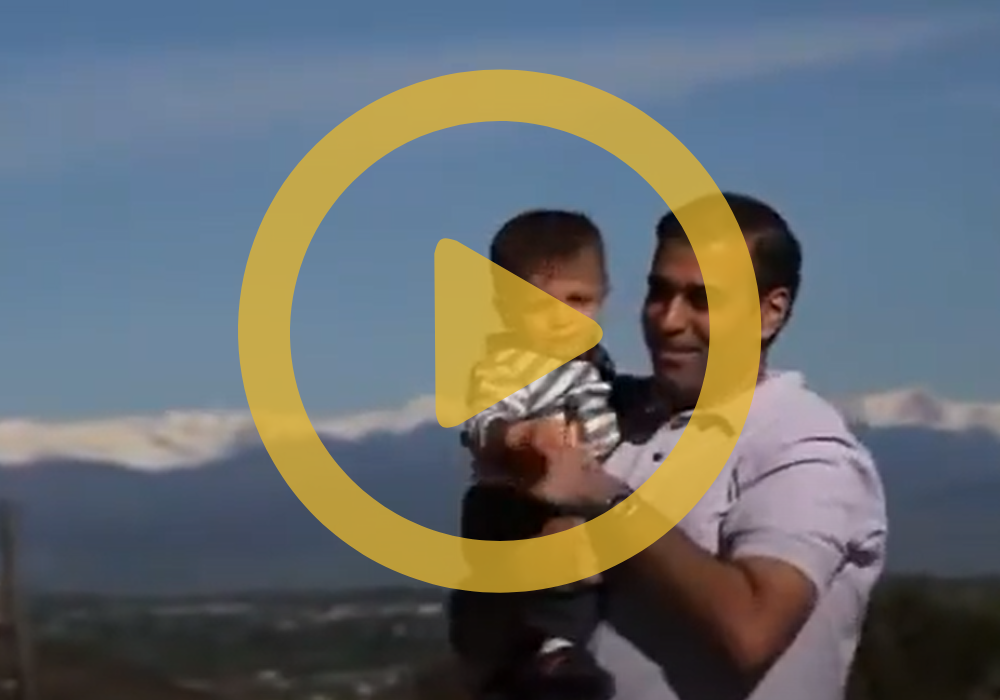 Dr. Shah holding his son with mountains in the background and a play button on the forefront