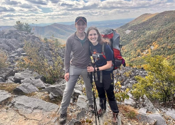 Two hikers stand on a rocky trail with scenic mountains in the background. Both are smiling and wearing hiking gear; one has a backpack and trekking poles. The landscape features autumn-colored foliage under a partly cloudy sky.