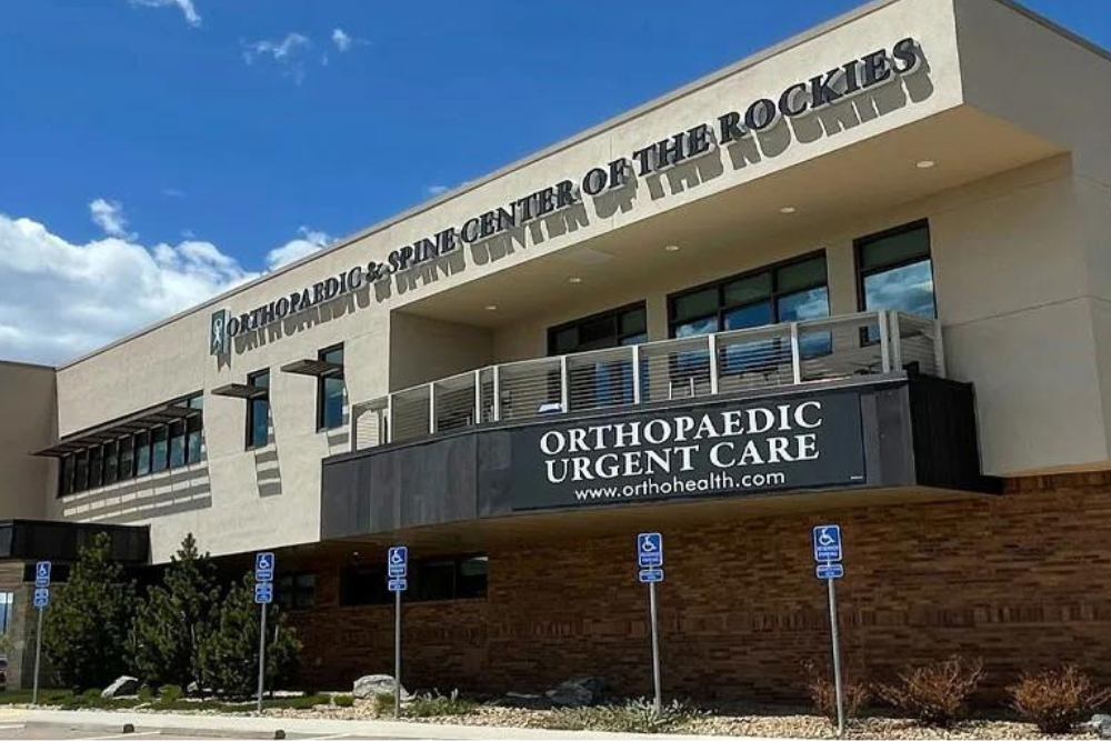 A two-story building with a sign "Orthopaedic & Spine Center of the Rockies" on the upper wall and "Orthopaedic Urgent Care" on a banner on the lower level. Expert care is provided for musculoskeletal injuries. There are several accessible parking spaces in front, and the sky is partly cloudy.