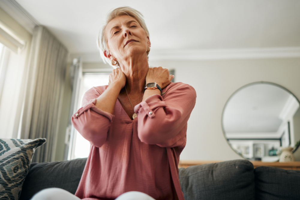 An older woman with short grey hair sits on a couch in a living room, holding her neck with both hands and appearing to be in discomfort, possibly pondering, "Will neck pain cause headaches?" She wears a pink blouse and a watch, with a round mirror visible on the wall behind her.