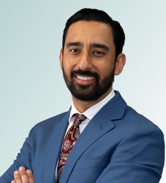 Patrik Suwak, a person with a beard, is smiling and wearing a blue suit jacket, a white dress shirt, and a patterned tie. He has dark hair and stands with his arms crossed against a light gradient background.