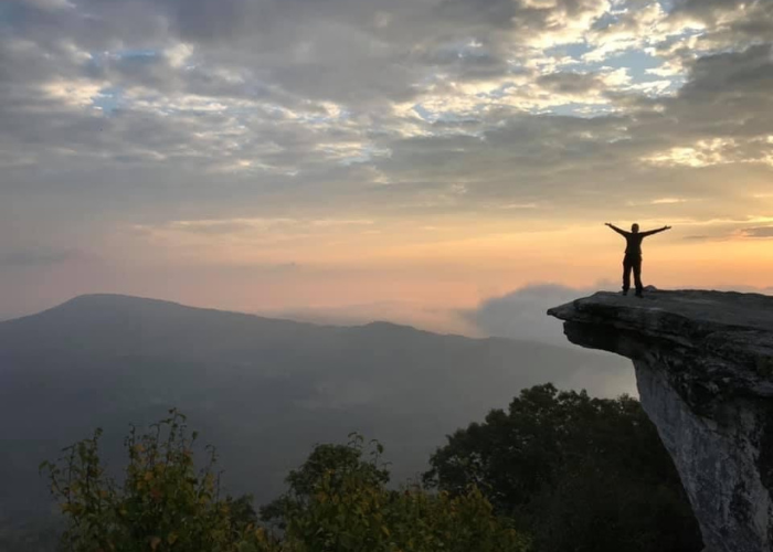 A person stands with arms outstretched on the edge of a rocky cliff overlooking a vast, mountainous landscape during sunrise or sunset. The sky is filled with clouds, casting a serene light over the scene. Vegetation can be seen in the foreground.