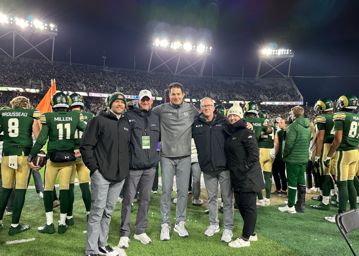 A group of five people stands on the sidelines of a football field at night, smiling for a photo. They are dressed warmly in jackets and hats. Behind them, football players in green and white uniforms, along with more people, are visible in front of illuminated stadium lights.