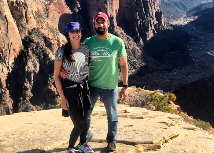 A couple stands on a rocky cliff with scenic canyon views in the background. The woman wears a blue cap and striped shirt, while the man wears a red cap and green t-shirt. They are both smiling and embracing each other amid the natural landscape.