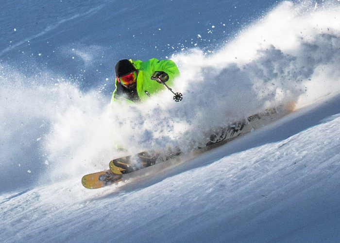 A skier dressed in a bright neon green jacket, black pants, and a black helmet is captured mid-action while skiing down a snowy slope. Snow sprays dramatically around them, highlighting the skier's dynamic movement through the fresh powder.