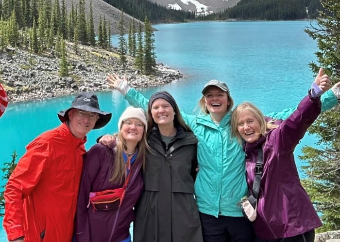 A group of five people, wearing colorful outdoor clothing, smile and pose cheerfully in front of a stunning turquoise lake surrounded by pine trees and mountains. The weather appears cool, and the group seems to be enjoying a nature adventure together.