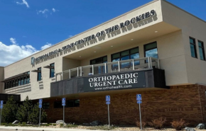 A two-story building with a sign that reads "Orthopaedic & Spine Center of the Rockies" and "Orthopaedic Urgent Care" on the front. The building has large windows on the second floor and blue sky with a few clouds in the background. Handicap parking spaces are visible.