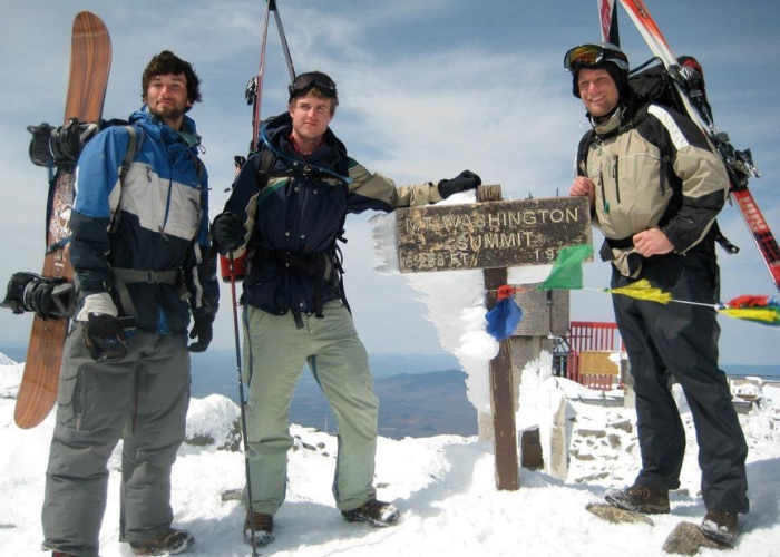 Three individuals dressed in winter gear stand at the snow-covered summit of a mountain next to a weathered sign that reads "Washington Summit" and other partially obscured text. Each person is holding skis or a snowboard. The sky is clear in the background.