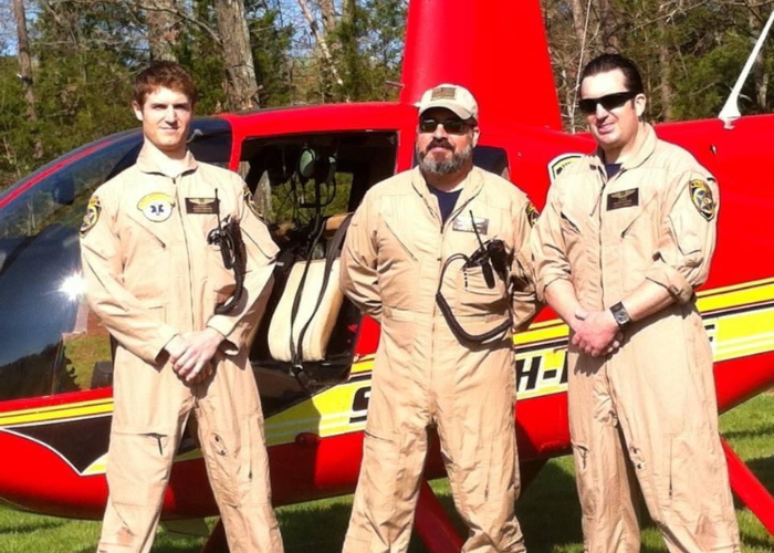 Three men in tan flight suits stand in front of a red helicopter with "SOUTH" partially visible on the side. The men have various gear attached to their suits and wear sunglasses. The background features trees and a grassy area.