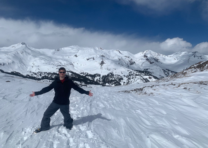 A person wearing sunglasses, a black jacket, and gray snow pants stands with outstretched arms on a snow-covered landscape with mountains in the background under a partly cloudy sky.