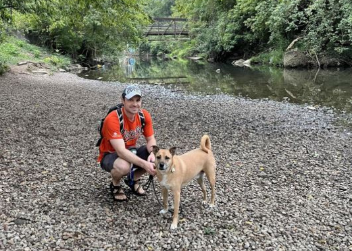 Dr. Johnson sits on a gravel trail in front of a river with his dog.