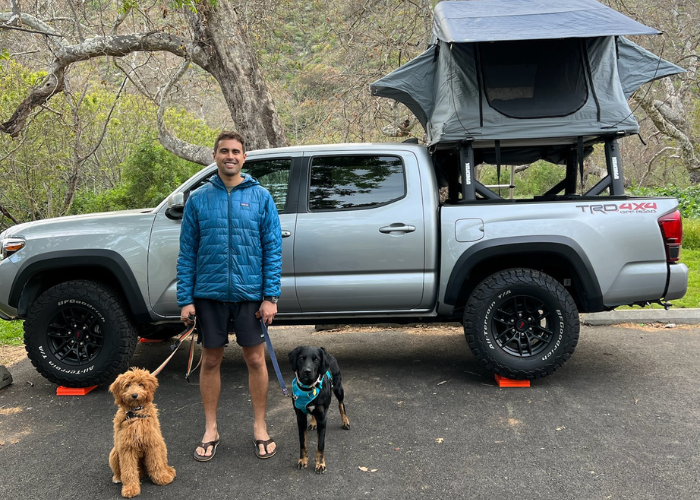 A man stands smiling next to a pickup truck with a rooftop tent. He holds leashes with two dogs: a fluffy brown dog on the left and a black dog with a blue bandana on the right. Trees and grassy areas are visible in the background.