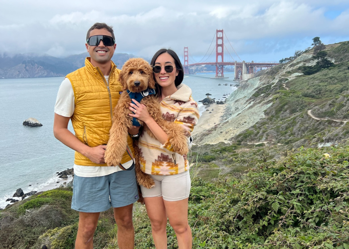 A couple stands on a scenic cliffside with the Golden Gate Bridge in the background. The woman holds a fluffy brown dog. Both are dressed in casual outdoor attire: the man in a yellow vest and shorts, the woman in a patterned jacket and shorts.