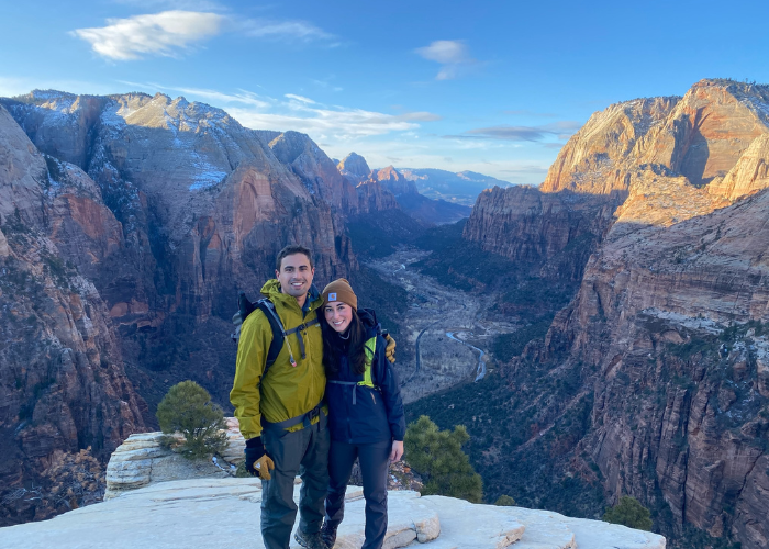 A man and woman stand together on a rocky ledge with a vast canyon landscape in the background. They are both wearing hiking gear, including jackets and beanies. The sun is setting, casting a warm glow on the mountains and the valley below.