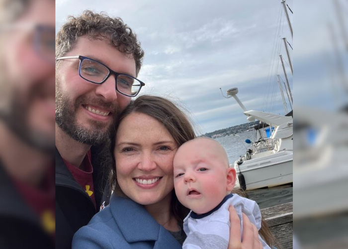 A man with glasses and a beard, a woman with long brown hair, and a baby are smiling in front of a marina with boats. The sky is cloudy. They appear to be enjoying a day out by the water.