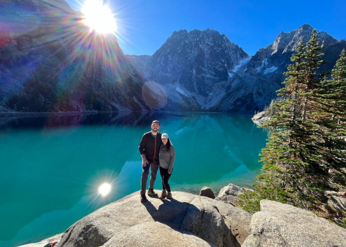 A man and a woman stand in front of a crystal blue lake with mountains and trees behind them.